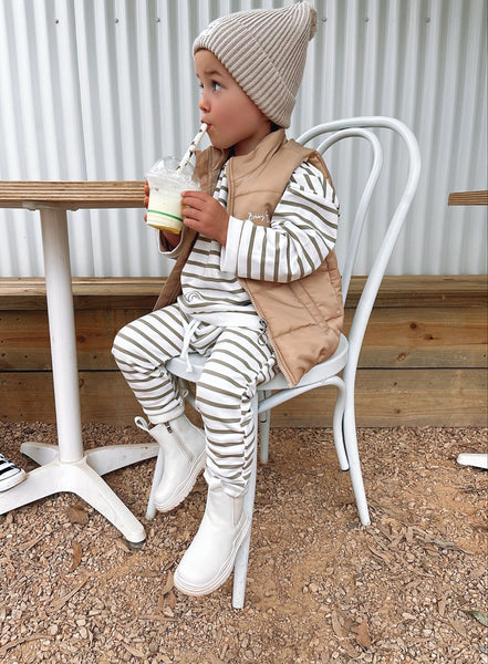 Little boy wearing a striped green and white organic cotton jumper and harem pants with a brown puffer vest that has a zip closure and Bobby G logo printed in white and a neutral knit beanie that has a Bobby G woven label attached 