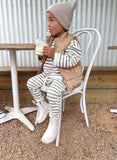 Toddler wearing oversized striped jumper and matching pants with a Winter vest jacket in brown and white Bobby G branding and neutral pom pom Bobby G beanie.  
