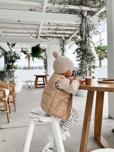 Toddler/child wearing neutral long sleeved waffle cotton top with organic cotton green and white striped pants and a kids winter brown vest with Bobby G printed on the chest in white and a neutral kids knit beanie with a pom pom and Bobby G woven label. 