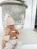 Child wearing neutral clothing, a waffle cotton long sleeved top with green and white striped pants and a brown vest jacket with Bobby G printed in white and neutral coloured knit beanie with Bobby G woven label attached.
